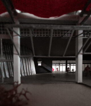 Blick aus dem Inneren eines Modellgebäudes mit Metallträgern, geschwungener Wand und Silhouetten von Miniaturfiguren durch einen großen offenen Eingang.