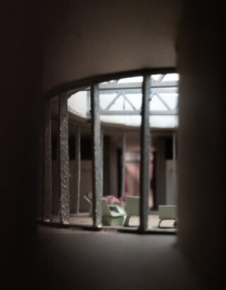 Blick durch vertikale Gitterstäbe in einen Raum mit einigen grünen Stühlen, großen Fenstern darüber und gedämpfter Beleuchtung, was an ein Architekturmodell oder einen Miniatur-Innenraum erinnert.