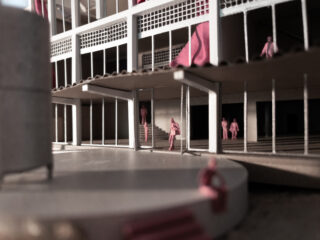 Architekturmodell, das ein mehrstöckiges Gebäude mit offenen Gehwegen, Treppen und kleinen rosafarbenen menschlichen Figuren zeigt, die um das Gebäude herum verteilt sind.