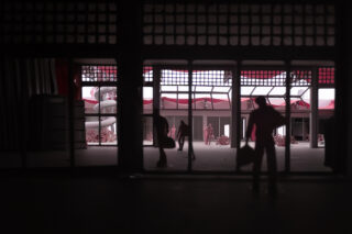 Ein dunkler Innenraum mit Silhouetten von Gestalten, die Taschen halten und durch eine gitterartige Wand und Glastüren ins helle Tageslicht blicken. Im sonnenbeschienenen Innenhof sind weitere Menschen zu sehen.