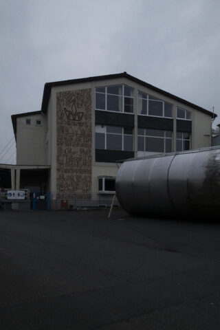 Ein zweistöckiges Gebäude mit großen Fenstern und einem Wandgemälde mit abstrakten Mustern an einer Wand, daneben eine große zylindrische Metallstruktur an einem bewölkten Tag.
