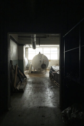 Ein schwach beleuchteter Industrieraum mit großen zylindrischen Tanks, gestapelten Paletten, Holzbrettern und Rohren in der Nähe eines Fensters.