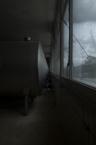 Großer Metalltank in einem schwach beleuchteten Raum neben einer Fensterreihe, durch das Glas ist die Landschaft im Freien sichtbar.