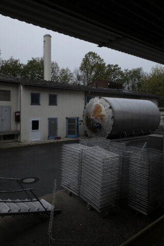 Ein großer zylindrischer Metalltank ist umgestürzt und liegt auf der Seite außerhalb eines Industriegebäudes, in der Nähe gestapelter Metallgestelle und einer gepflasterten Fläche.