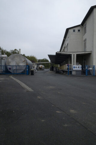 Industrieanlage mit großen zylindrischen Tanks, einem mehrstöckigen Gebäude, blauen Metalltoren und einer gepflasterten Auffahrt unter einem bewölkten Himmel.