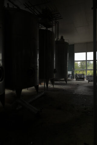 Große Industrietanks stehen in einem schwach beleuchteten Raum mit Rohren auf dem Boden; Sonnenlicht fällt durch Fenster im Hintergrund ein.