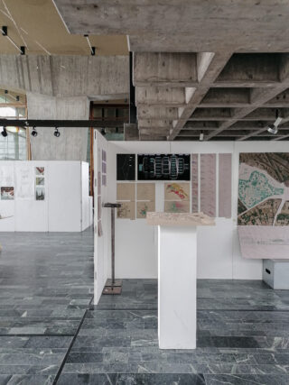 Ein architektonischer Ausstellungsraum mit Schautafeln mit Entwurfsskizzen, Renderings und Karten in einem modernen Betoninterieur mit hoher Decke.