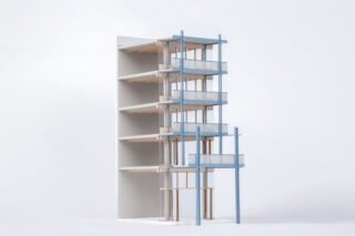 Ein minimalistisches Architekturmodell eines fünfstöckigen Gebäudeteils mit freiliegenden Holzböden und blauen Strukturstützen auf der rechten Seite vor einem schlichten weißen Hintergrund.