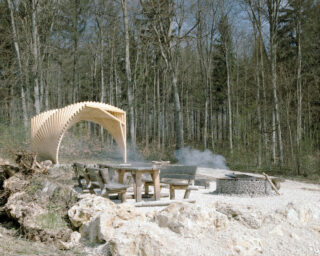Sitzbereich im Freien mit Holzbänken und -tischen, einem geschwungenen Holzdach und einer runden, rauchenden Feuerstelle auf einer Waldlichtung.