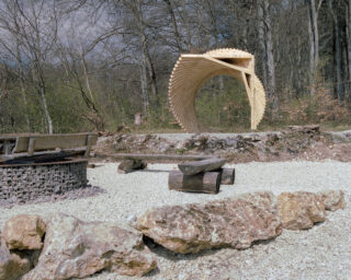 Eine spiralförmige Holzskulptur steht auf einer Kieslichtung in der Nähe einer Feuerstelle aus Stein und einer rustikalen Sitzgelegenheit aus Holzstämmen, umgeben von blattlosen Bäumen und Felsen.