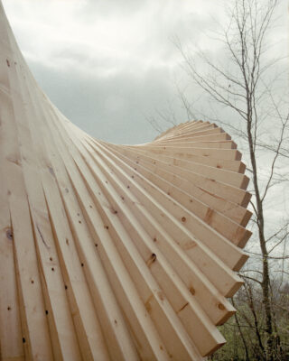 Eine Nahaufnahme einer modernen architektonischen Holzstruktur mit einem gewundenen, gerippten Design, die im Freien in der Nähe blattloser Bäume unter einem bewölkten Himmel steht.