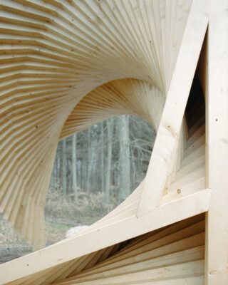 Nahaufnahme einer Holzkonstruktion mit eckigen, überlappenden Paneelen, die vor einem Hintergrund aus Bäumen ein verdrehtes Spiralmuster bilden.