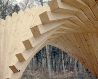 Nahaufnahme einer hölzernen Architekturstruktur mit überlappenden Balken, die einen abgewinkelten Bogen bilden, im Freien mit Bäumen im Hintergrund.