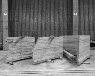 Stapel von Holzbrettern sind in drei Gruppen auf einem Betonboden in einem großen Industrieraum mit holzgetäfelten Wänden angeordnet.