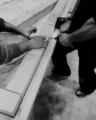 Zwei Personen bauen mithilfe einer Bohrmaschine eine Holzkonstruktion zusammen, wobei sie die Bretter mit den Händen auf einer Werkbank halten und führen.