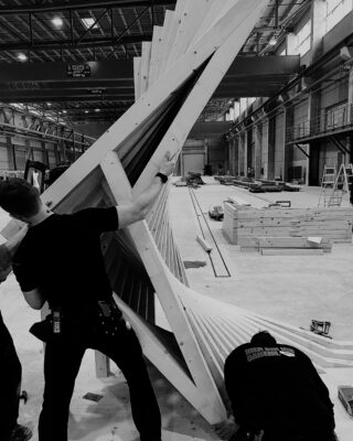 Zwei Arbeiter montieren in einer geräumigen Industriehalle eine große, verdrehte Holzkonstruktion.