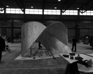 Große Holzskulptur mit ineinandergreifenden gebogenen Latten steht in einer geräumigen Industriewerkstatt; im Hintergrund sind Menschen und Werkzeuge zu sehen.
