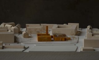 Ein maßstabsgetreues Architekturmodell mit weißen Gebäuden zeigt von oben betrachtet eine zentrale Struktur in Orange, umgeben von Miniaturbäumen und Straßen.