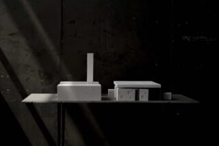 Auf einem Tisch sind mehrere rechteckige und quadratische Betonblöcke unterschiedlicher Größe ausgestellt, die vor einer dunklen Wand mit diagonalen Lichtstreifen angeordnet sind.