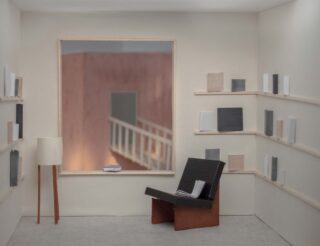 Ein minimalistischer Raum mit weißen Wänden, Büchern auf schmalen Regalen, einem schwarzen Stuhl mit einem offenen Buch, einer Stehlampe und einem großen Fenster mit Blick auf ein Geländer und eine Treppe.