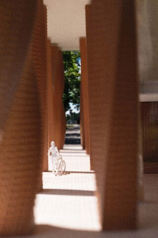 Eine kleine weiße Figur mit einem Fahrrad steht zwischen hohen, strukturierten Säulen unter einem überdachten Gehweg, im Hintergrund sind Sonnenlicht und Grün zu sehen.