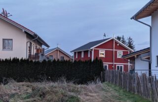 Ein rotes zweistöckiges Haus mit weißen Verzierungen steht zwischen zwei modernen Häusern, getrennt durch eine hohe Hecke und einen Holzzaun in einem Vorortviertel.