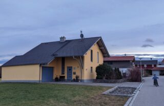 Neben einer ruhigen Straße steht ein gelbes Haus mit grauem Dach. Im Vordergrund ist ein Junge zu sehen, der auf dem Bürgersteig Fahrrad fährt.