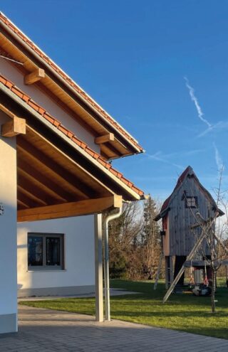 Im Vordergrund ist ein weißes Haus mit rotem Ziegeldach zu sehen. Im Hintergrund steht ein hölzernes Baumhaus auf Stelzen in einem grasbewachsenen Hof unter einem klaren blauen Himmel.