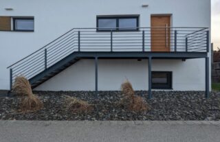 Moderne Hausfassade mit einer Metalltreppe und einem Geländer, die zu einer Holztür über einem mit Kies bedeckten Boden und trockenen Ziergräsern führen.