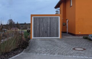 Neben einem orangefarbenen Haus steht eine freistehende Holzgarage mit geschlossenem Tor, im Vordergrund eine gepflasterte Auffahrt und einige kahle Sträucher.