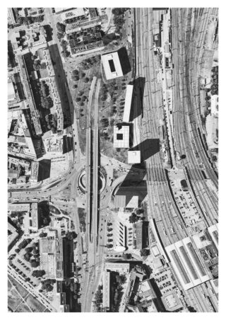 Luftaufnahme eines Stadtgebiets mit Straßen, einem Kreisverkehr, Gebäuden und Bahngleisen mit Bahnsteigen auf der rechten Seite.