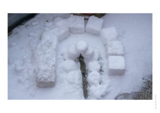 Eine kleine Schneestruktur auf dem Boden, bestehend aus bogenförmig angeordneten Schneeblöcken und gestapelten Schneebällen, die einen Bogen bilden, umgeben von Schnee.