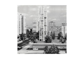 Ein moderner Glasturm steht zwischen Wohnhäusern aus der Mitte des Jahrhunderts in einem Stadtgebiet. Im Vordergrund sind Bäume, geparkte Autos und ein Zebrastreifen zu sehen.