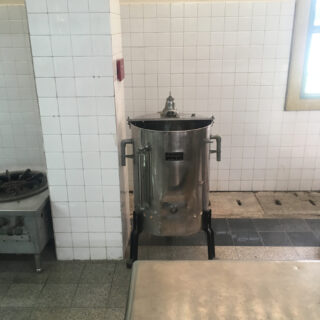 An einer gefliesten Wand in einer Großküche steht ein großer Schnellkochtopf aus Edelstahl, links davon ein Gasherd und im Vordergrund ein Tisch.