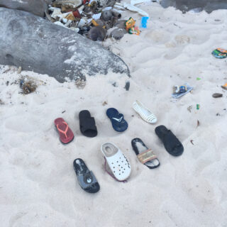 Acht nicht zueinander passende, weggeworfene Sandalen und Flip-Flops liegen in einer Reihe am Sandstrand in der Nähe von Felsen und diversem Müll.