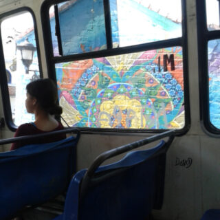 Eine Frau sitzt allein in einem Bus und blickt aus dem Fenster auf ein farbenfrohes geometrisches Wandgemälde, das draußen auf eine Backsteinmauer gemalt ist.