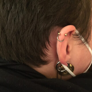 Nahaufnahme des Ohrs einer Person mit mehreren Piercings, darunter ein goldener Industrial Bar, ein Creolen-Piercing und ein großer Plug. Die Person trägt eine Gesichtsmaske mit weißen Bändern.