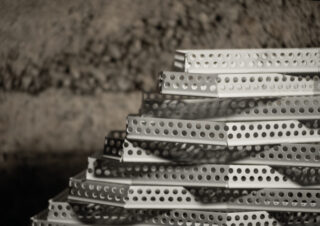 Ein Stapel silberner perforierter Metallstreifen, pyramidenförmig angeordnet, mit einer rauen Steinmauer im Hintergrund.