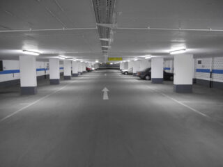 Eine Tiefgarage mit heller Deckenbeleuchtung, weißen Säulen und einigen an den Seiten geparkten Autos; auf den Boden ist ein weißer Pfeil gemalt, der die Richtung anzeigt.