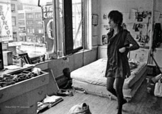 Eine Frau steht in einem überfüllten, sonnendurchfluteten Schlafzimmer mit einem ungemachten Bett und einem Blick durch das Fenster auf eine Stadtstraße.