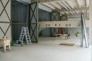 Innenansicht eines unfertigen Industriegebäudes mit Leitern, Tischen, Balken, einer Palette und verstreuten Baumaterialien.