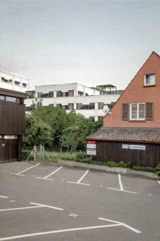 Ein leerer Parkplatz vor einem braunen Holzgebäude und einem orangefarbenen Haus, mit modernen weißen Gebäuden und Grün im Hintergrund.