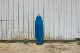 Ein blaues Surfbrett steht aufrecht vor einer Wellblechwand, an dessen Fuß auf einer Betonoberfläche ein paar kleine Unkräuter wachsen.