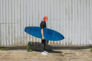 Eine Person mit roter Mütze, schwarzem Hemd und weißen Turnschuhen steht seitlich und hält ein blaues Surfbrett vor einer Wellblechwand.