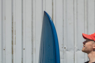 Ein Mann mit einer roten Mütze blickt neben der Spitze eines blauen Surfbretts nach oben, das an einer Wellblechwand steht.