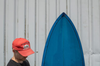 Eine Person mit einer roten Marlboro-Kappe steht neben der Spitze eines blauen Surfbretts vor einer Wellblechwand.