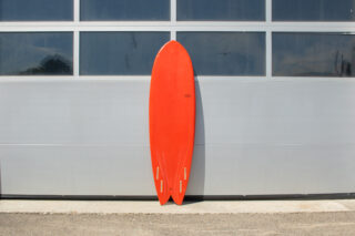 Ein rotes Surfbrett im Fischstil mit vier Flossen steht aufrecht vor einer grauen Metallwand mit großen Fenstern.