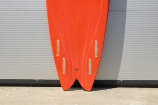 Der untere Teil eines roten Surfbretts mit Schwalbenschwanz-Design und vier Finnenkästen steht aufrecht vor einer Wellblechwand.