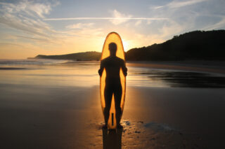 Eine Person steht an einem Sandstrand und hält ein Surfbrett aufrecht. Die Silhouette zeichnet sich vor dem Sonnenuntergang mit Hügeln und Wolken im Hintergrund ab.