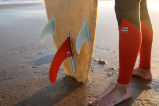Eine Person im Neoprenanzug steht barfuß auf nassem Sand neben einem Surfbrett mit drei Finnen, einer roten und zwei weißen, die teilweise im Strand vergraben sind.